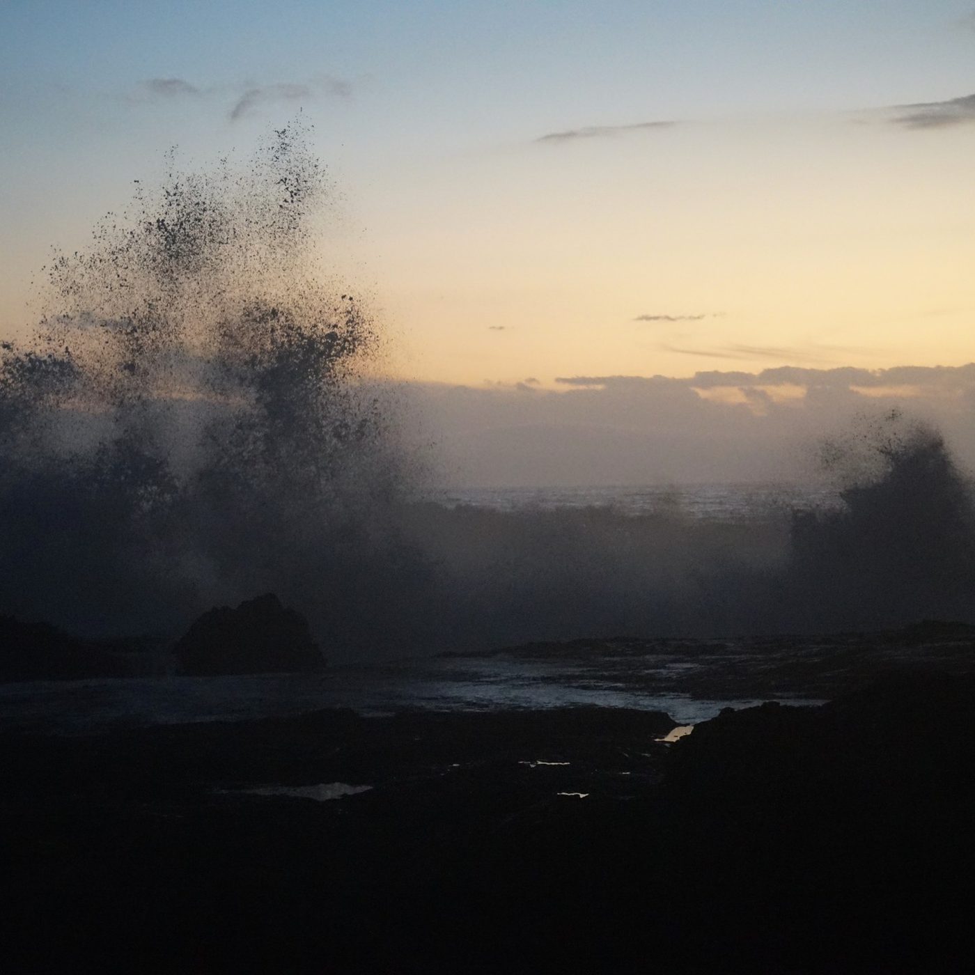 Waves breaking over dark rocks at dusk, spray thrown high against the sky